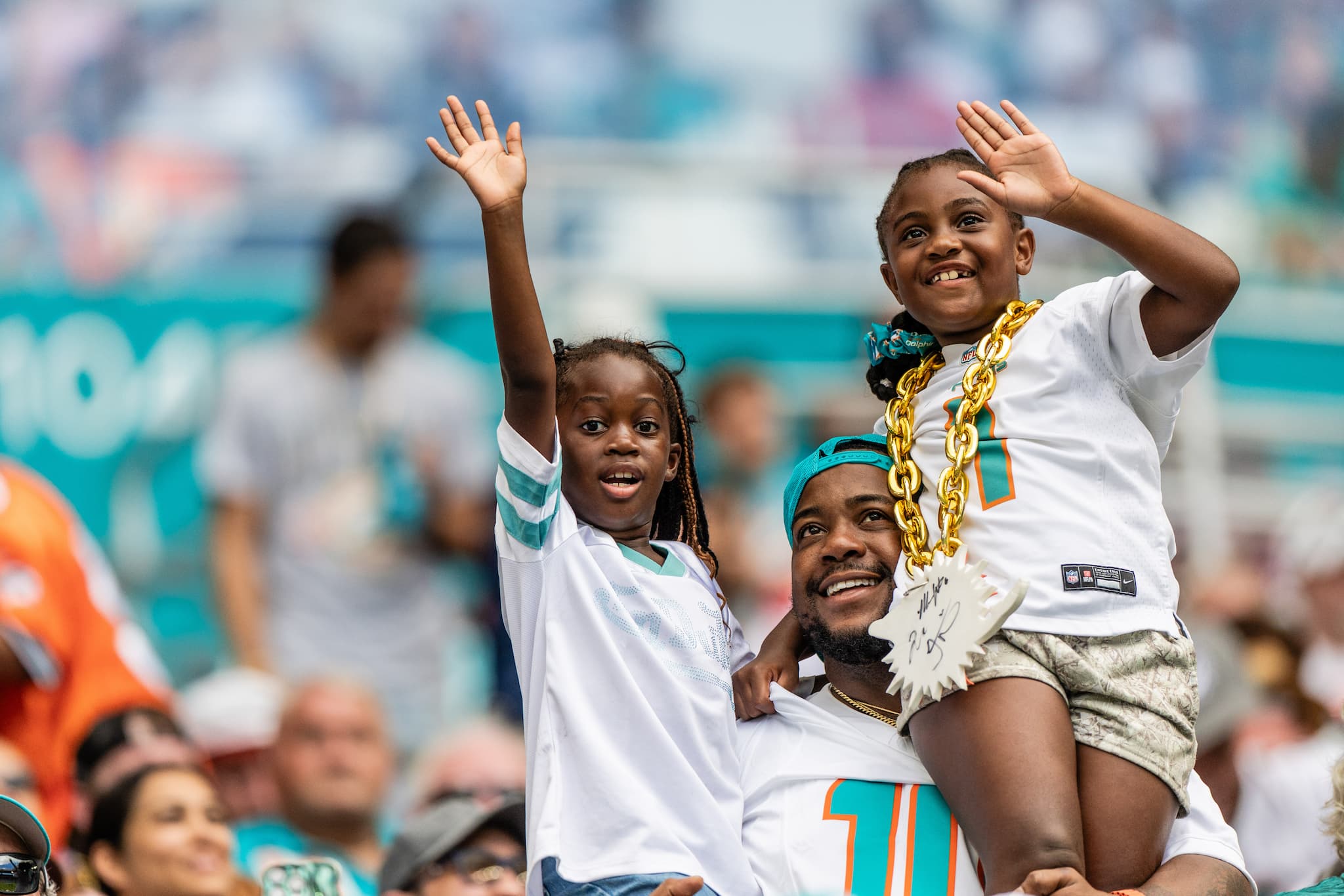 f475cf7bee3b2b43bf17dd51400b5e6fd8247199-2 A father holds his two children up to wave at the camera in a game, all decked in Dolphins gear.